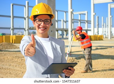 Happy beautiful female architect with tablet on construction site. She is smiling and satisfied with her job, giving thumb up looking at camera, behind her construction worker with measuring device