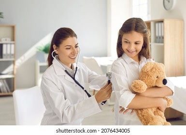 Happy beautiful female doctor holding stethoscope and doing lungs exam to little child. Smiling pediatrician checking her patient's respiration during routine checkup at modern pediatric clinic