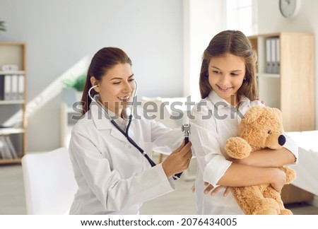 Happy beautiful female doctor holding stethoscope and doing lungs exam to little child. Smiling pediatrician checking her patient's respiration during routine checkup at modern pediatric clinic