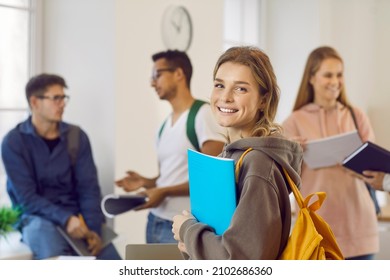 Happy Beautiful Female University Student With Book And Backpack. Portrait Of Pretty Young Girl With Candid Cheerful Face Expression Smiling At Camera While Standing In Classroom With Her Classmates