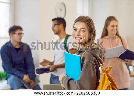 Happy beautiful female university student with book and backpack. Portrait of pretty young girl with candid cheerful face expression smiling at camera while standing in classroom with her classmates