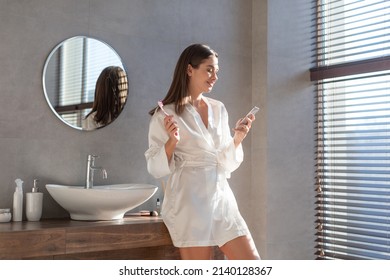 Happy Beautiful Female Usign Smartphone While Brushing Teeth In Bathroom In The Morning, Young Woman Enjoying Online Communication Or Internet Shopping While Making Daily Hygiene At Home, Copy Space - Powered by Shutterstock