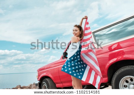 happy beautiful girl with american flag standing near red car