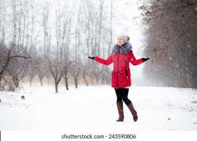 Happy beautiful girl in knitted hat and red winter coat, outdoors in park, rejoicing at snowfall, full length
