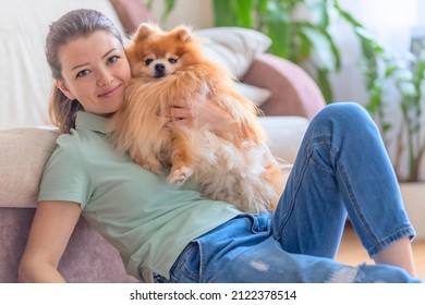 happy beautiful girl playing with dog at home. pomeranian playing with owner. love to animals. human pet friendship, adoption. young cute woman hugging spitz
