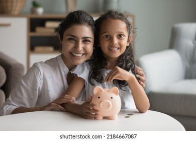 Happy beautiful Indian mother and kid girl saving money, holding piggy bank, looking at camera, smiling, laughing. Mom and child making reserve fund, investment, donations. Home portrait