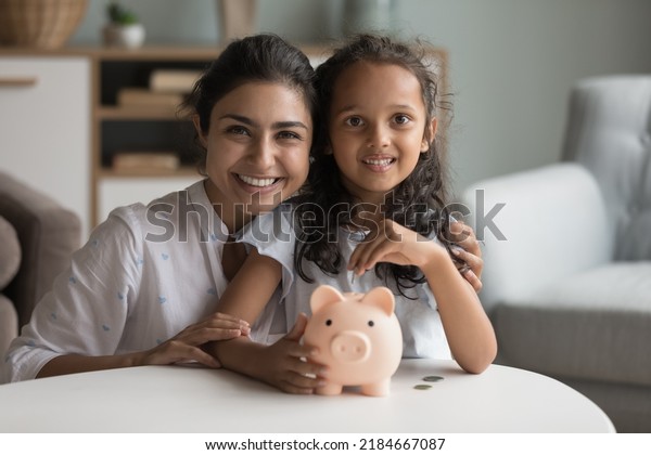 Happy\
beautiful Indian mother and kid girl saving money, holding piggy\
bank, looking at camera, smiling, laughing. Mom and child making\
reserve fund, investment, donations. Home\
portrait