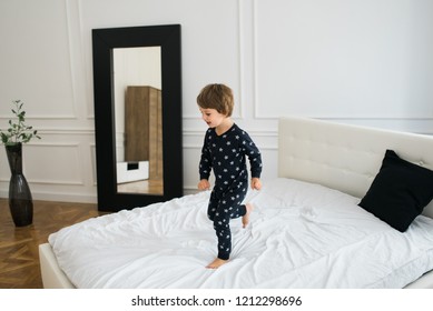 Happy Beautiful Little Boy Jumping On The Bed Early In The Morning Wearing One Piece Pajamas.