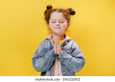 Happy beautiful little girl child join hands pray with hope meditating, isolated on yellow studio background wall, calm religious small kid feel gratitude thanking in prayer, faith meditation concept