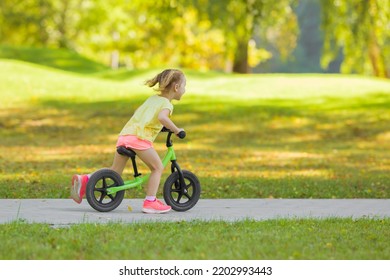 Happy Beautiful Little Girl Fast Running And Riding On First Bike Without Pedals On Sidewalk At City Park In Warm Summer Day. Cute 3 Years Old Toddler. Side View. Learning To Keep Balance.