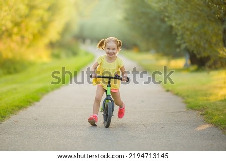 Happy beautiful little girl fast running and riding on first bike without pedals on sidewalk at city park in warm summer day. Cute 3 years old toddler. Front view. Learning to keep balance.