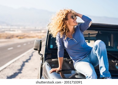 Happy Beautiful Mature Woman In Sunglasses Posing With Hand In Curly Hair While Sitting On Bonnet Of Jeep At Highway. Cheerful Stylish Woman Posing On Car Bonnet At Roadside During Her Road Trip