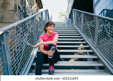 Happy And Beautiful Middle Aged Woman Sitting On Metallic Stairs Relaxing Before Running Outdoors Holding A Water Bottle