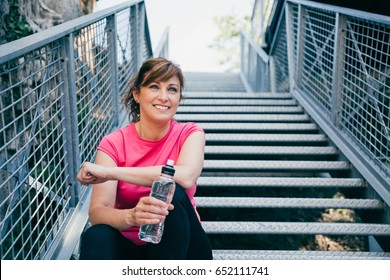 Happy and beautiful middle aged woman sitting on metallic stairs relaxing before running outdoors holding a water bottle