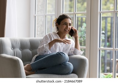 Happy beautiful millennial Indian woman talking on mobile phone call, enjoying conversation, communication, leisure, resting on soft armchair at window, smiling, speaking. Wireless connection - Shutterstock ID 2184667089