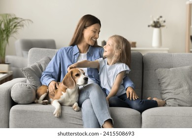 Happy beautiful mother embracing kid and touching dog on home sofa, talking to little daughter girl, smiling, laughing, relaxing in cozy home interior, spending family leisure with kid and pet - Powered by Shutterstock