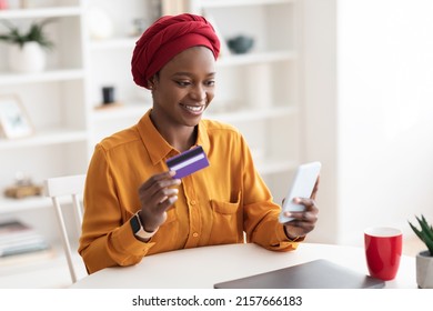 Happy beautiful muslim millennial african american woman with sitting at workdesk, using brand new smartphone and credit card, buying goods and services on Internet, banking online, copy space - Powered by Shutterstock