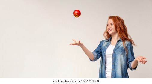 Happy beautiful redhead girl throwing apple in the air on light studio background, panorama with copy space
