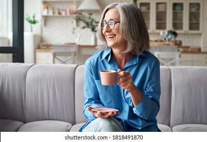 Happy beautiful relaxed mature older adult grey-haired woman drinking coffee relaxing on sofa at home. Smiling stylish middle aged 60s lady enjoying resting sitting on couch in modern living room.