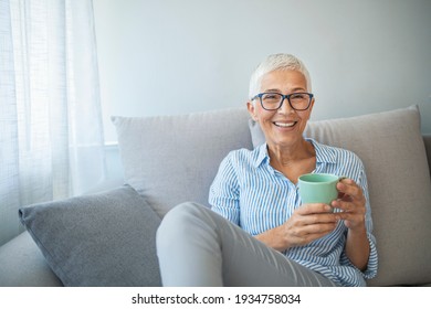 Happy beautiful relaxed mature older adult grey-haired woman drinking coffee relaxing on sofa at home. Smiling stylish middle aged 60s lady enjoying resting sitting on couch in modern living room. - Powered by Shutterstock