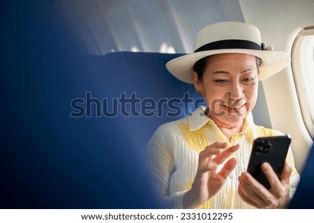 A happy and beautiful senior Asian female passenger using her smartphone at her window seat on a flight, traveling for her summer trip. tourist, vacation, airplane mode
