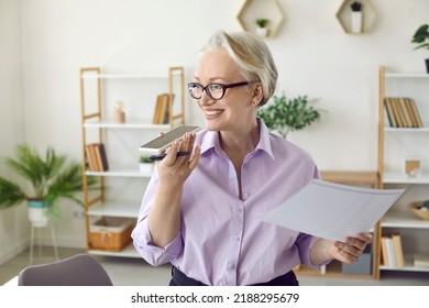 Happy beautiful senior business woman holding mobile phone and documents, smiling, talking on speaker phone, recording voice message, using Internet search virtual assistant or audio note taking app - Powered by Shutterstock