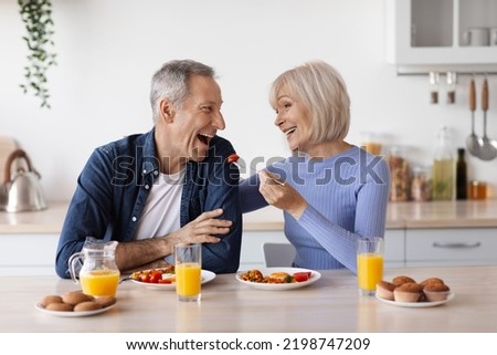 Happy beautiful senior woman having healthy lunch with her husband at home, elderly spouses sitting at kitchen table, eating vegetables and having conversation, feeding each other, copy space