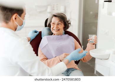 Happy beautiful senior woman sitting in dental chair, holding glass of water, talking with dentist doctor after dental treatment. Concept of service, dental care - Powered by Shutterstock