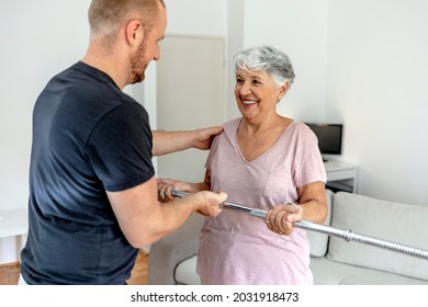 Happy Beautiful Senior Woman Smiling And Holding Easy Bar While Exercising With Personal Trainer. An Older Woman Lifting Weights In Rehabilitation Center. Elderly Lady Is Exercising With Man At Home