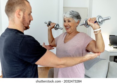 Happy Beautiful Senior Woman Smiling And Holding Easy Bar While Exercising With Personal Trainer. An Older Woman Lifting Weights In Rehabilitation Center. Elderly Lady Is Exercising With Man At Home