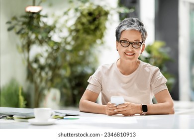 Happy beautiful short-haired middle aged woman in smart casual and eyeglasses manager enjoying coffee break, sitting at workplace office coworking space, using smartphone, sending email, copy space - Shutterstock ID 2346810913