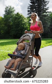 Happy Beautiful Sports Mom In A Pink Top And Black Leggings Runs A Morning Jogging With A Small Baby With A Pacifier Sitting In A Pram In A Summer Sunny Park