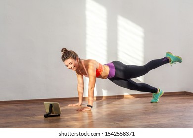 Happy beautiful sporty woman with bun hairstyle and in tight sportswear doing plank with leg raise while watching training video on tablet. indoor studio shot illuminated by sunlight from window - Powered by Shutterstock