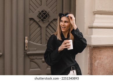 Happy beautiful stylish woman having good fashion clothes walking on street and holding coffee in cup takeaway with good mood. Girl look happy, look at side, put on or put off glasses, hold it by hand