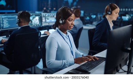 Happy Beautiful Technical Customer Support Specialist Is Talking On A Headset While Working On A Computer In A Call Center Control Room Filled With Colleagues, Display Screens And Data Servers.