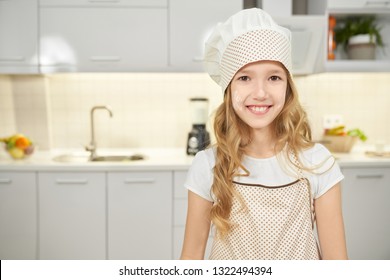 Happy beautiful teenager standing in kitchen, looking at camera and smiling. Charming girl wearing in apron and white chef hat. Cheerful kid posing, having flour on face.