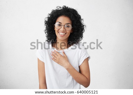 Happy beautiful warm-hearted young dark-skinned female wearing big round spectacles and white t-shirt smiling and holding hand on her breast while watching touching romantic scene in TV series