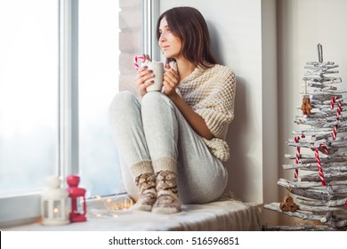 Happy Beautiful Woman Drinking Hot Coffee Sitting On Window Sill In Christmas Decorated Home. Holiday Concept 