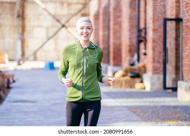 Happy Beautiful Woman Jogging Outdoor. Healthy Female Slim Running On The City Street. Middle Aged Lady In Activewear Running With Red Brick Industrial Building In Background.