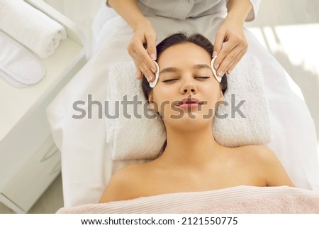 Happy beautiful woman lying on spa massage bed in beauty parlor and enjoying modern facial treatment. Beautician removing makeup with cotton discs and cleaning skin before rejuvenating detox procedure ストックフォト © 