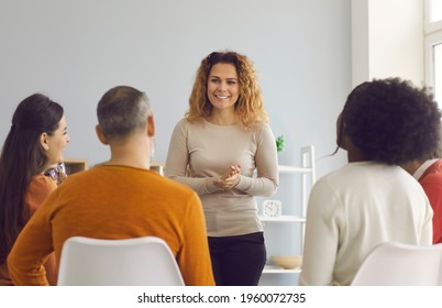 Happy Beautiful Woman Making Presentation Standing In Front Of Diverse Audience. Psychologist Or Coach Talking To Group Of Young And Mature People. Team Of Company Workers Listening To Female Speaker