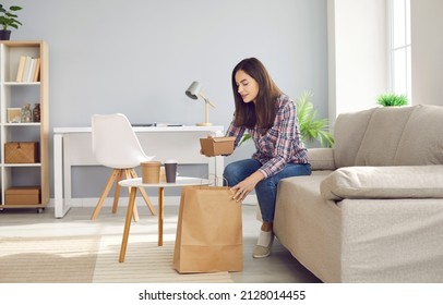 Happy Beautiful Woman Opens Paper Bag With Her Takeaway Meal. Young Girl Sitting On Sofa At Home And Unpacking Containers With Healthy Takeout Food. Food Delivery Concept