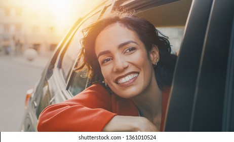 Happy Beautiful Woman Riding On A Back Seat Of A Car, Looks Out Of The Open Window In Wonder. Traveling Girl Experience Magic Of The World. Sunny Shot Made From Outside The Vehicle.