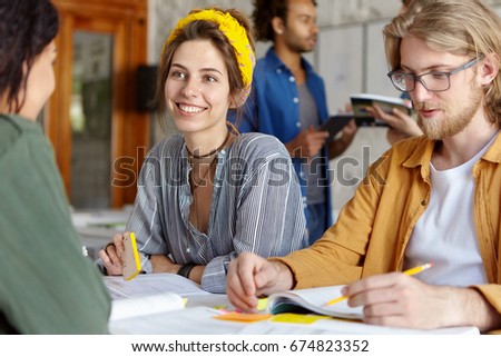 Happy beautiful woman in shirt discussing something with her African friend sitting near her groupmate with beard. Team of young people working together using books. Communication and team concept