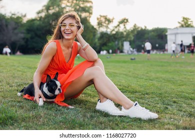 Happy Beautiful Woman Sitting On Grass In Summer Park, Holding Boston Terrier Dog, Smiling Positive Mood, Wearing Orange Dress, Trendy Style, Slim Legs, Sneakers, Playing With Pet