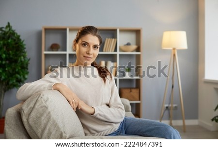 Happy beautiful woman sitting on the couch at home and looking at the camera. Portrait of a young girl in her 20s or 30s relaxing on her comfortable sofa in her modern living room