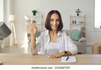 Happy Beautiful Woman Sitting At Work Desk With Notebook And Pen At Home, Waving Hello And Smiling. Web Cam Laptop Computer Screen View Of Adult Student In Online Lecture, Videocall Or Videoconference