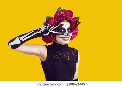 Happy beautiful woman with skull face makeup and flowers on head does peace victory sign with two fingers, smiles and looks at camera isolated on yellow background. Halloween, Day of the Dead concept - Shutterstock ID 2198491449