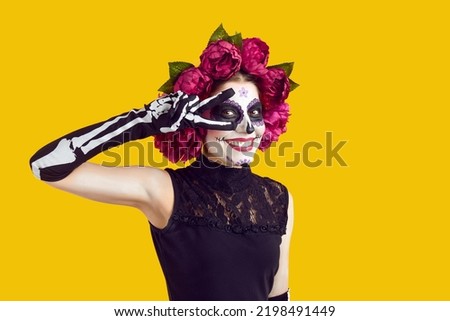 Happy beautiful woman with skull face makeup and flowers on head does peace victory sign with two fingers, smiles and looks at camera isolated on yellow background. Halloween, Day of the Dead concept