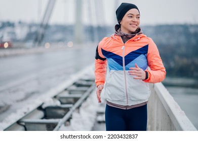 Happy Beautiful Woman In Sportswear And A Black Cap And Running On The Bridge At Winter. Healthy Lifestyle, Winter Fitness, Cold Weather. Copy Space. Girl Trains Endurance.